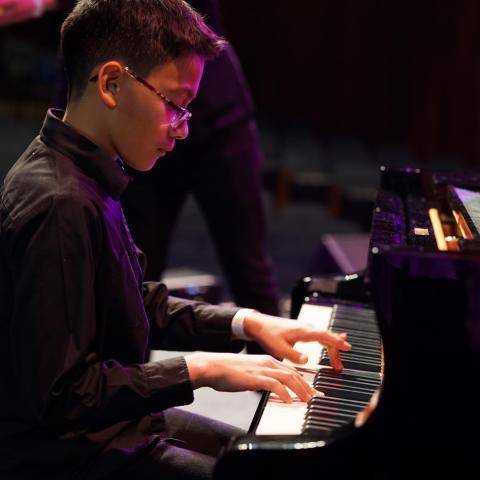 Preparatory Academy student with dark hair and glasses is seated at an upright piano at the Berklee Performance Center.